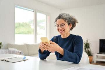 Happy cheerful older woman customer using smartphone sitting at home table in living room. Smiling middle aged lady holding cellphone doing shopping buying in smartphone app, texting messages on phone
