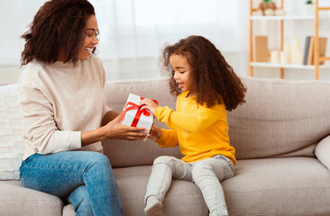 Family Holiday. Happy Afro Mother Congratulating Little Daughter Giving Her Birthday Gift Sitting On Sofa At Home.