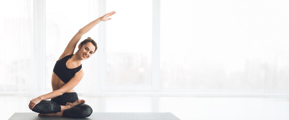 A woman performs a side stretch in a modern yoga studio filled with natural light, panorama with...