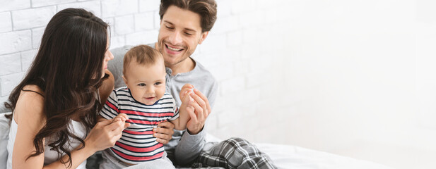 A joyful family is enjoying quality time together in their bedroom. The mother and father are playfully interacting with their young child, who is smiling and engaging with them, copy space