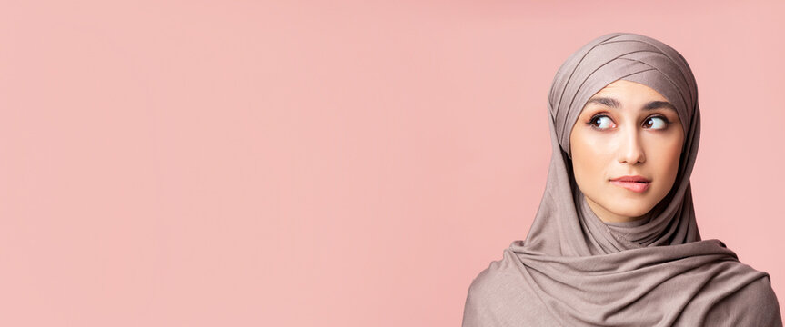 Closeup portrait of uncertain muslim girl in hijab biting lip and looking away at copy space over pink background, having doubts about something