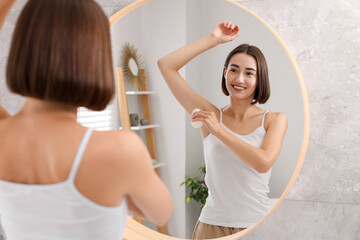 Smiling woman applying roll-on deodorant near mirror at home, back view