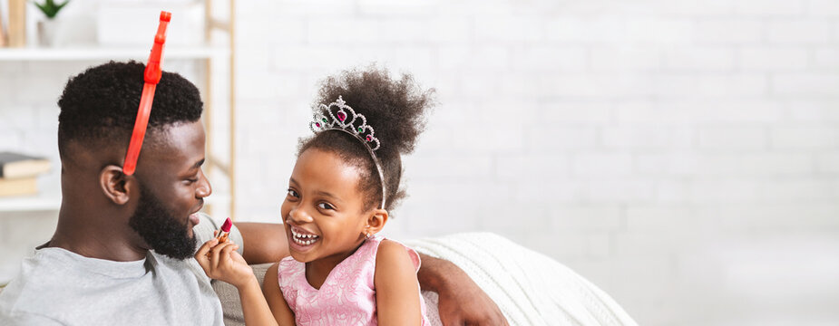 Black father and his daughter share a delightful moment, surrounded by light and warmth in their living room. The little girl wears a playful tiara and smiles brightly as she engages with her dad