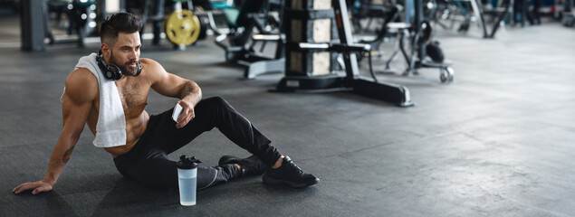 A young man sits on the gym floor, taking a break after exercising, with a towel around his neck and a water bottle nearby, copy space