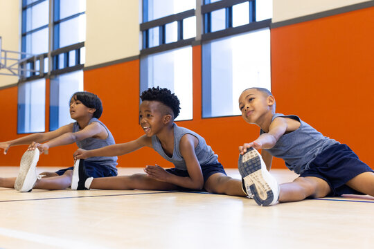 In school gym, three multiracial boys stretching on floor, smiling and exercising together