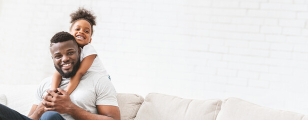 A joyful black father interacts with his young daughter, who playfully hugs him from behind while they sit on a comfortable sofa. The warm atmosphere highlights their close bond, copy space