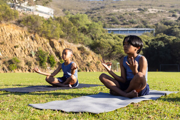 Practicing yoga, two multiracial boys meditating on mats outdoors at school field