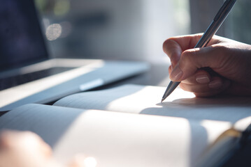 Close up of woman hand with a pen writing on notebook with laptop computer on office table....