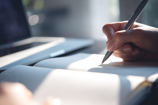 Close up of woman hand with a pen writing on notebook with laptop computer on office table. Business woman planning, making a note reminder. Student studying online, e-learning