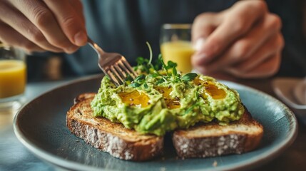 A soft focus image shows a man enjoying a hearty and delectable breakfast in a hip café or restaurant. He is ready to indulge and satisfy his hunger by spreading avocado spread or guacamole over rye b