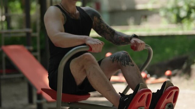 A young man exercising outdoors on a public rowing machine. He is wearing a black tank top, black shorts, and a black baseball cap. He is working out on a bright sunny day.