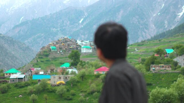 4K Rack focus shot of an Indian man looking at Triloknath village in front of the snowy Himalayan mountain peaks at Pattan valley in Lahaul, Himachal Pradesh, India. Man looking at the village.