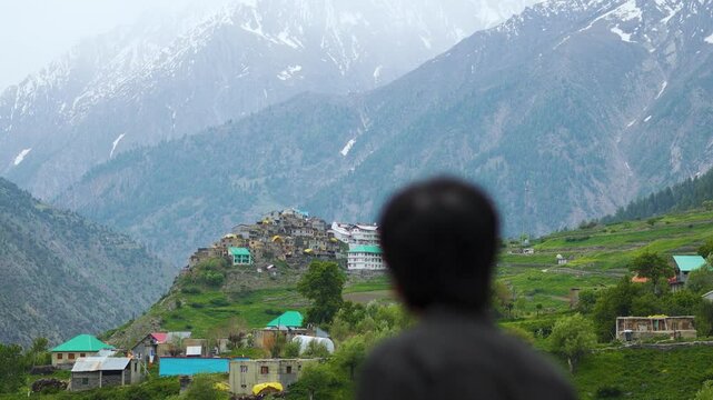 4K shot of a defocused Indian man looking at Triloknath village in front of the snowy Himalayan mountain peaks at Pattan valley in Lahaul, Himachal Pradesh, India. Man looking at the village on cliff.