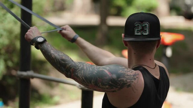 A fit man with tattoos wearing a black cap and a black tank top doing tricep extensions using a resistance band on an outdoor calisthenics gym