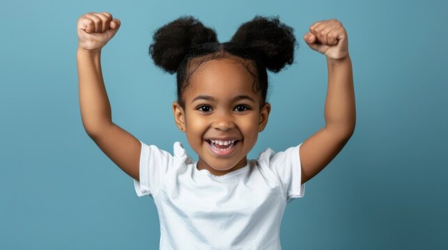 A little Black girl with a big smile and her fists raised in the air celebrates a victory or achievement.&nbsp;She is wearing a white shirt and her dark hair is styled in a high ponytail,&nbsp;copy space