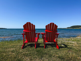 Two red adirondack chairs on grass overlooking ocean on sunny day. © Cavan