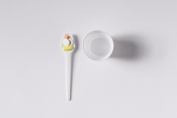 Plastic spoon with pills and cup of water on white background