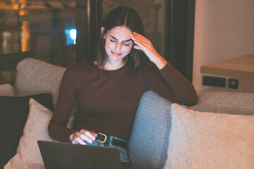 A woman sitting on the sofa at home late at night looking disappointed and distressed while using a...