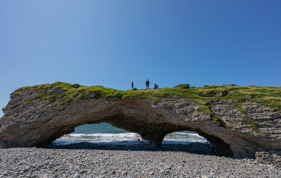 Three people atop natural rock arches on beach in Newfoundland.