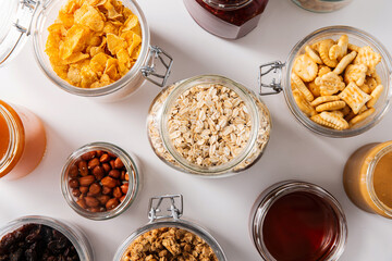 food storage and eating concept - close up of jars with oat, corn flakes, granola, cookies and spreads on white background