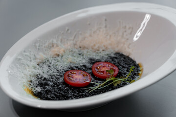 Gourmet Culinary Presentation of Elegant Black Risotto Garnished with Tomato Slices and Herbs in a White Bowl on a Sophisticated Gray Backdrop