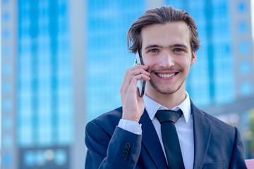 Smiling businessman near a business center calling by cell phone. A happy young businessman talking with a smartphone. Young cheerful businessman near a modern tall glass building with a phone. .