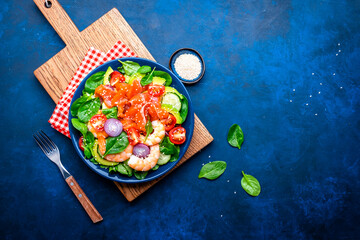 Healthy food salmon salad for keto diet with shrimp, avocado, spinach, cucumber, tomato, cashew, sesame seeds. Blue background, top view