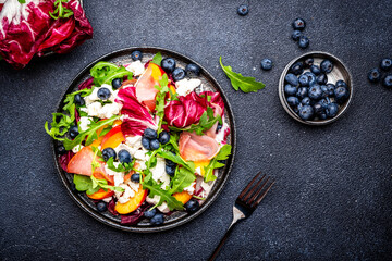 Foodie food salad with peach, jamon, soft cheese, blueberries and arugula on black background, top view