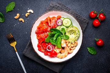 Seafood salad bowl with salmon, shrimp, avocado, spinach, cucumber, tomato, cashew and sesame. Black background, top view
