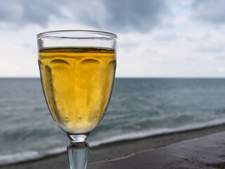 A glass filled with chilled white wine stands against a backdrop of the ocean. The tranquil waves and overcast sky create a calming, relaxing atmosphere.