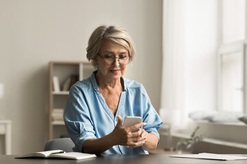 Digital communication, tasks management using mobile application. Senior woman sitting at desk at home holding smartphone, making call, ordering through virtual e-services, buying, browsing internet