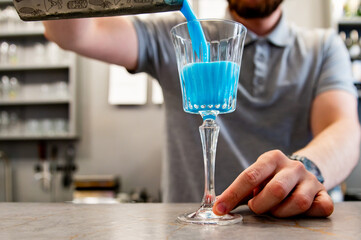 A bartender pours a vibrant blue cocktail into a glass on a bar counter, capturing the dynamic motion of the liquid. The scene is set in a stylish bar environment, emphasizing the elegance drink.
