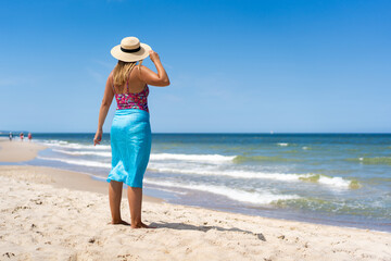 Summer vacation. Tanned woman in colorful swimsuit, blue pareo and sun hat standing by shore on sandy beach on beautiful sunny day. Back view	