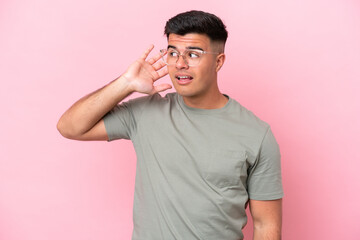 Young caucasian handsome man isolated on pink background listening to something by putting hand on the ear