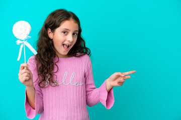 Little caucasian girl holding a lollipop isolated on blue background surprised and pointing finger to the side