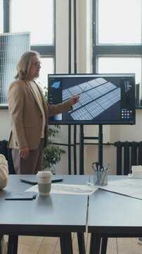 Full vertical shot of middle-aged male engineer presenting new solar generation project on multimedia screen to colleagues at team meeting in office of renewable energy startup