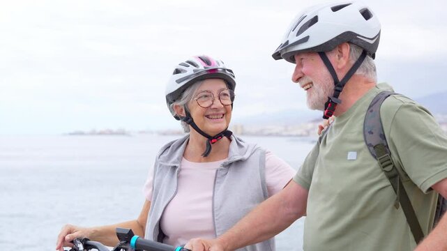Senior couple riding bikes together in rocky beach enjoying outdoor. Active mature people talking holding mountain bikes. Bicycle lifestyle concept, having fun
