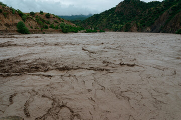 Rushing muddy river water is carrying sediment and debris downstream after heavy rain in a mountain valley. The power of nature is visible in the muddy water