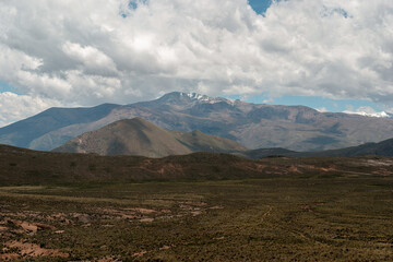 The andes mountains dominate the horizon, their snow-capped peaks reaching towards a sky filled with dramatic clouds casting shadows on the barren landscape