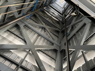 A low-angle view inside a tall industrial structure shows metal beams and girders forming a framework, with cables, wires, and an elevator shaft visible. A glimpse of the sky is seen at the top. © Yojang