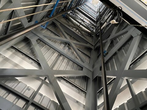 A low-angle view inside a tall industrial structure shows metal beams and girders forming a framework, with cables, wires, and an elevator shaft visible. A glimpse of the sky is seen at the top.