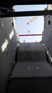 A large, red metal beam is being lifted by a yellow crane into a large, corrugated metal structure. The crane's long boom extends high into the blue sky, dotted with fluffy white clouds.