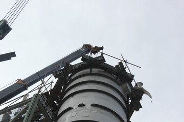 A large industrial chimney or silo is being constructed or repaired. A crane with a hook and scaffolding are visible against a cloudy sky. The focus is on the top of the structure with metal beams  