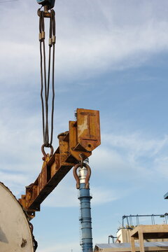 A large, rusty metal beam is being lifted by a crane against a blue sky with white clouds. The beam has hooks attached to it and is suspended by thick cables. An industrial factory with chimneys  
