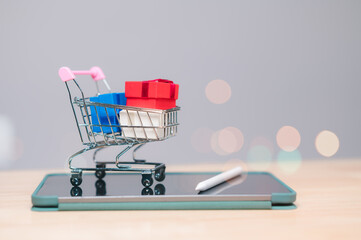 A miniature shopping cart filled with colorful gift boxes sits on a tablet, symbolizing the...