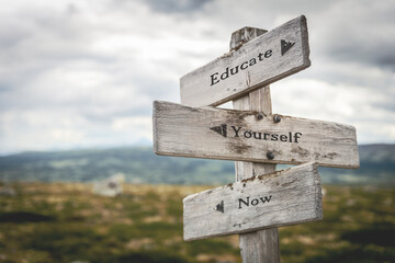 educate yourself now text quote on wooden directional signpost outdoors in nature