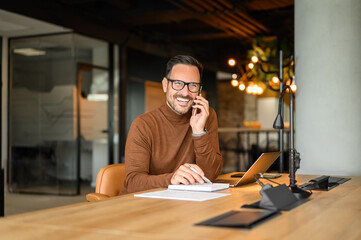 Happy adult businessman discussing over phone call and looking away while working at desk in office