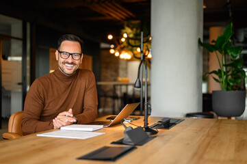 Portrait of inspired male entrepreneur in glasses working over laptop while sitting at desk in...