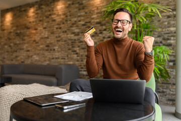 Satisfied businessman with credit card and laptop screaming and celebrating reward at table in...