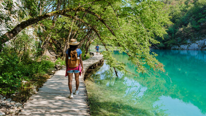 Amidst the lush greenery, travelers stroll along the wooden walkways of Plitvice Lakes National...